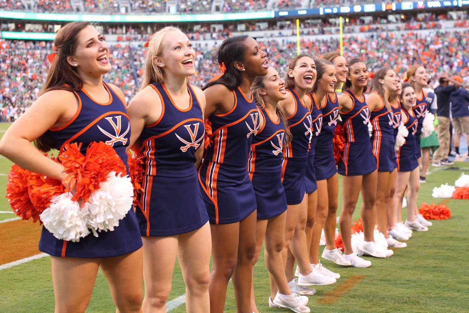 Virginia Cavaliers cheerleaders linking arms in celebration after a touchdown against the Notre Dame Fighting Irish at Scott Stadium in Charlottesville, VA, on September 12, 2018. Dressed in navy and orange uniforms, they stand on the sidelines, smiling and cheering as the crowd erupts in excitement. The stadium lights illuminate the scene, highlighting the energy of the moment.