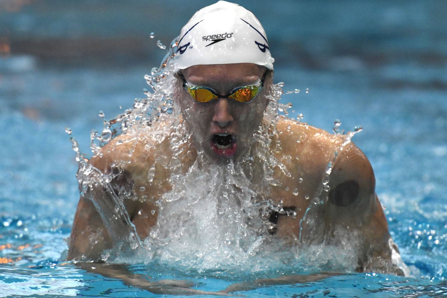 Jared Norton of the Virginia Cavaliers competing in the men's 100-yard breaststroke at the Aquatic Fitness Center in Charlottesville, VA, on January 14, 2017. Wearing a navy swim cap with the Virginia logo, he powers through the water with strong strokes, creating ripples around him. The pool's lane markers and overhead lighting frame the action, emphasizing the competitive atmosphere of the race.