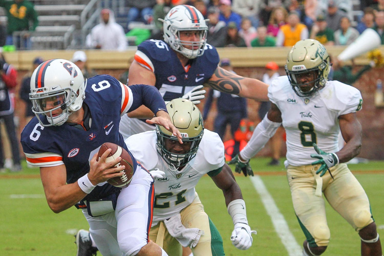 Kurt Benkert (#6), quarterback for the Virginia Cavaliers, evading two William & Mary defenders during a football game at Scott Stadium in Charlottesville, VA, on September 9, 2017. Benkert holds the football tightly as he maneuvers past the defenders, showcasing his agility and determination. The stadium lights illuminate the field, adding to the intensity of the moment.