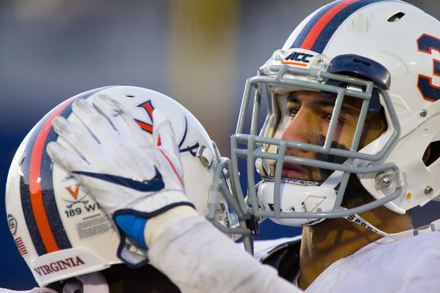Quin Blanding (#3) of the Virginia Cavaliers shares an emotional embrace with a teammate in the final moments of the Military Bowl at Navy-Marine Corps Memorial Stadium in Annapolis, MD, on December 28, 2017. The two players, dressed in white and navy uniforms, stand near the sideline, their expressions reflecting the intensity and emotion of the game. The stadium lights cast a glow over the scene, emphasizing the significance of the moment.