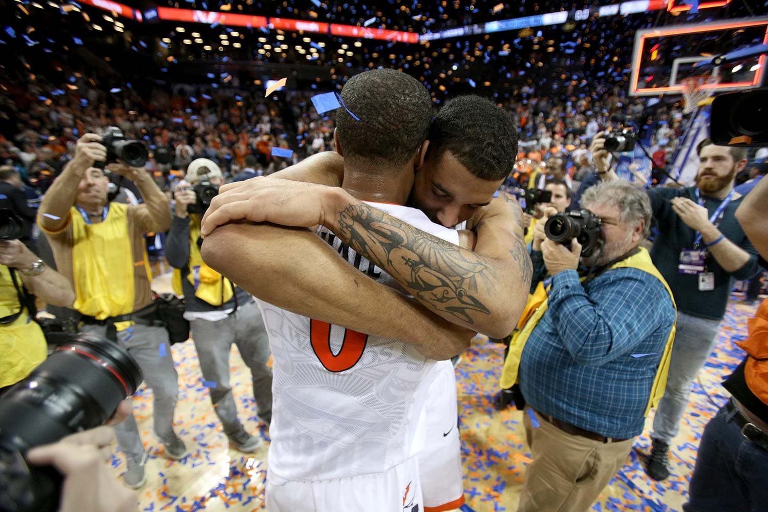 Teammates Devon Hall (0) and Isaiah Wilkins (21) of the Virginia Cavaliers celebrating their victory over the North Carolina Tar Heels in the ACC Championship game. The players are embracing each other, with Hall holding the championship trophy aloft. The Barclays Center is filled with cheering fans and falling confetti, creating a vibrant and triumphant atmosphere.