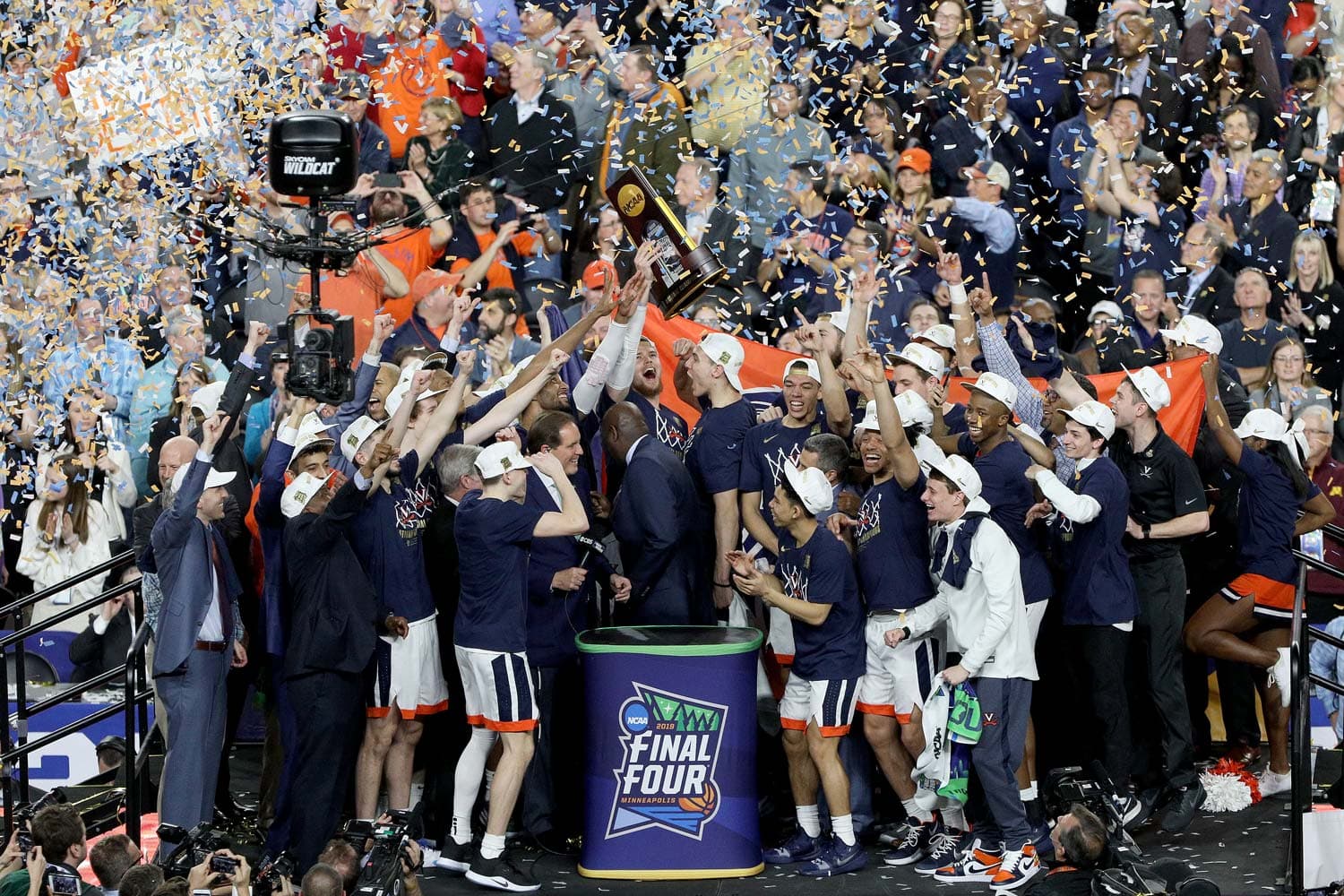 Virginia Cavaliers basketball team celebrating their NCAA national championship victory. Players and coaches gather around the podium, holding up the championship trophy as confetti falls from above. The crowd cheers in the background, capturing the excitement and triumph of the moment.