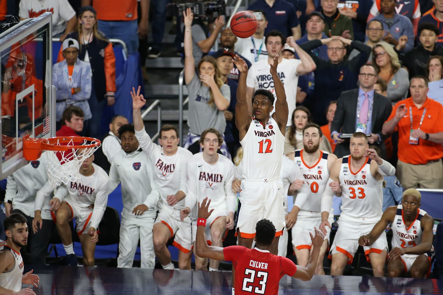 De'Andre Hunter of the Virginia Cavaliers, wearing jersey number 12, captured mid-air taking a shot during the 2019 National Championship game. The basketball is visible above his head, with the hoop positioned to the left. Texas Tech’s Jarrett Culver, wearing jersey number 23, is seen in the foreground with arms raised, attempting to block the shot. The Virginia Cavaliers bench is in the background, with players and staff watching intently.