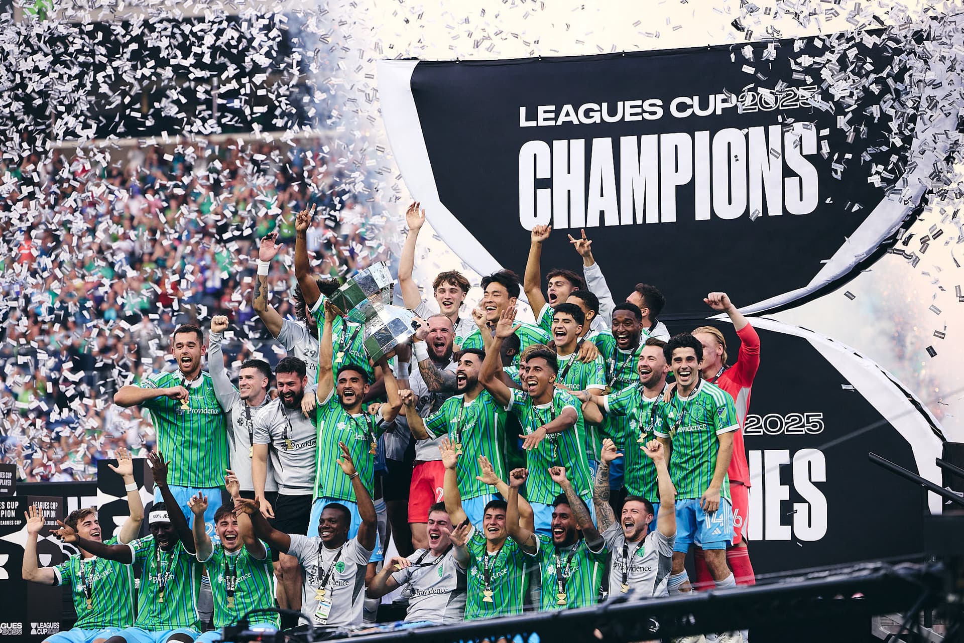 Seattle Sounders FC players celebrate on stage after winning the Leagues Cup Final at Lumen Field. Fireworks explode in the background as confetti rains down on the team.
