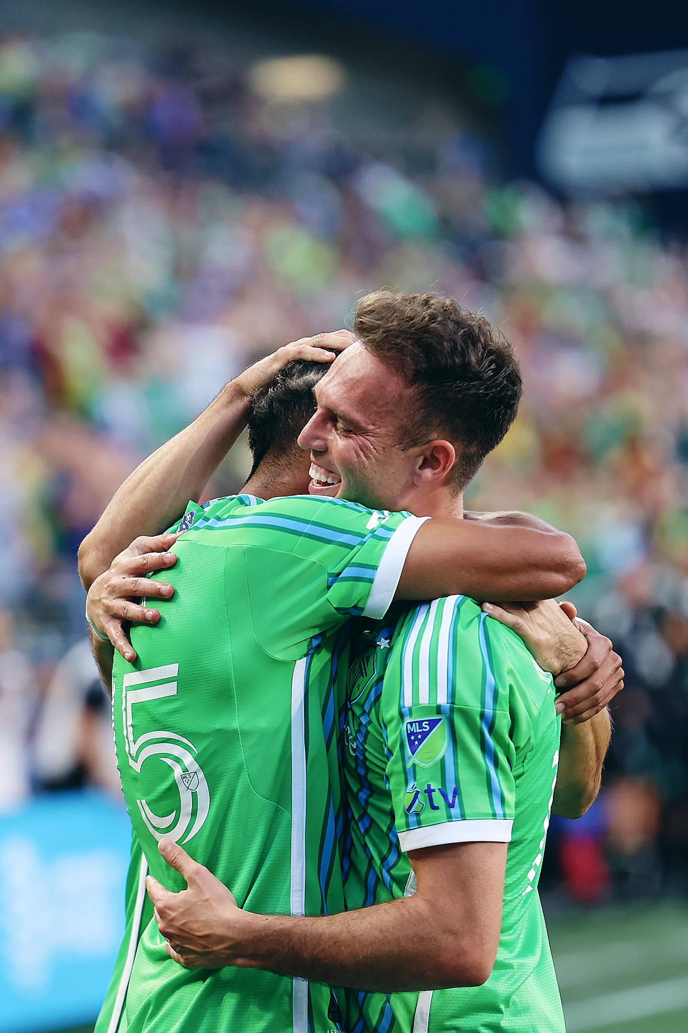 Danny Musovski of Seattle Sounders FC celebrates after scoring a goal against Sporting Kansas City during the second half of an MLS match at Lumen Field. Musovski is seen in embrace with his teammates, wearing the team's green and blue kit.