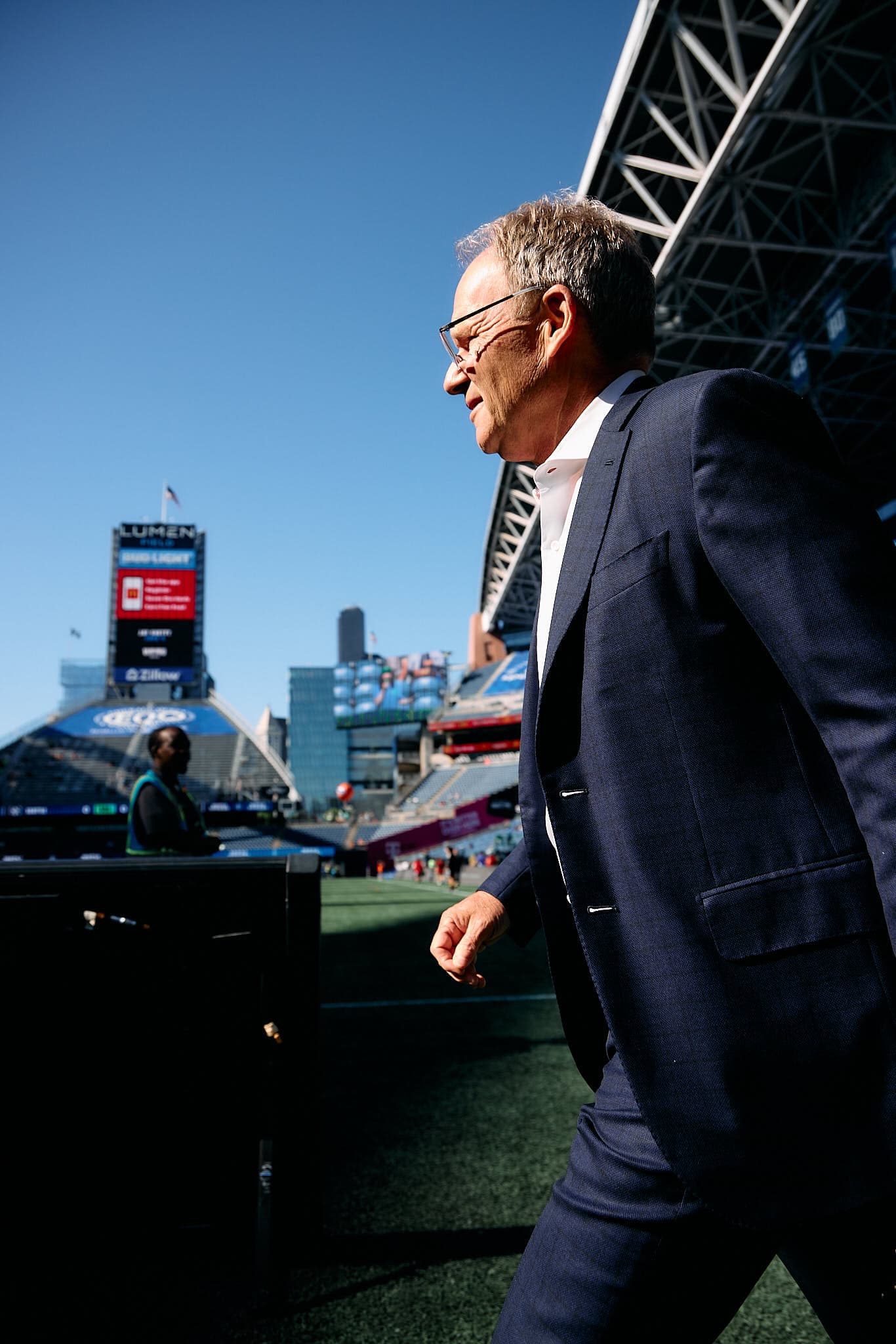 Brian Schmetzer, head coach of the Seattle Sounders FC, is captured walking across the field during a Major League Soccer match. He is dressed in a dark jacket and pants, with a focused expression as he prepares for the game. The vibrant green field and the stadium's stands filled with fans create an energetic backdrop, emphasizing the anticipation of the match.