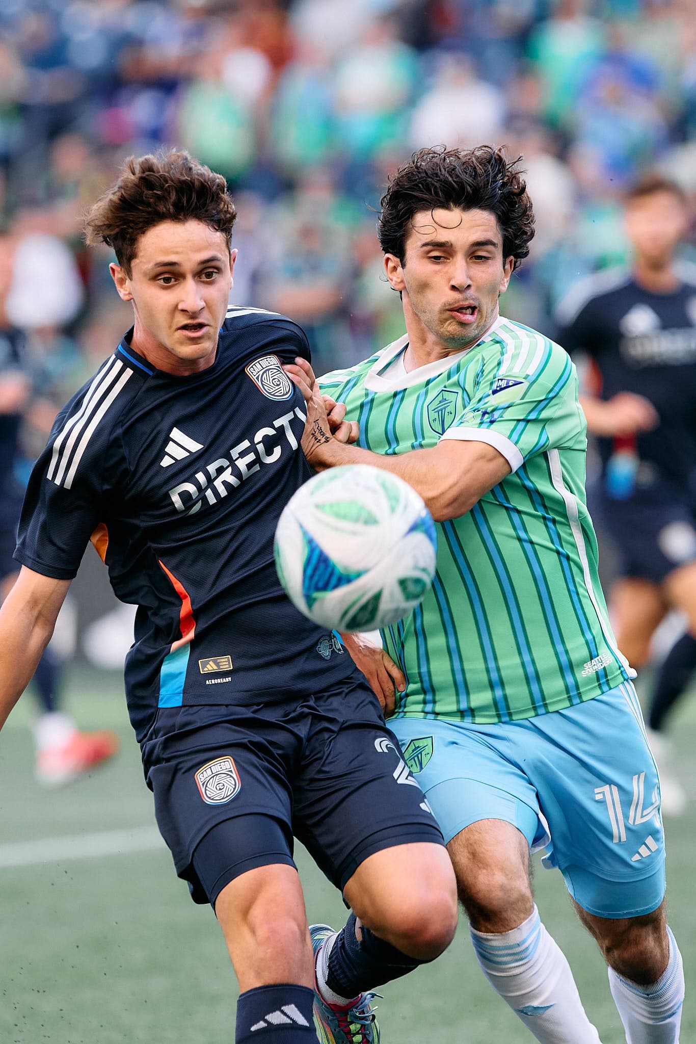 Paul Rothrock of the Seattle Sounders FC, wearing jersey number 14, is seen in a dynamic moment during a Major League Soccer match. He is engaged in a physical challenge for the ball against San Diego FC's Luca Bombino, who wears jersey number 27. The image captures the intensity of the game, with both players focused on the ball, surrounded by a vibrant stadium atmosphere.