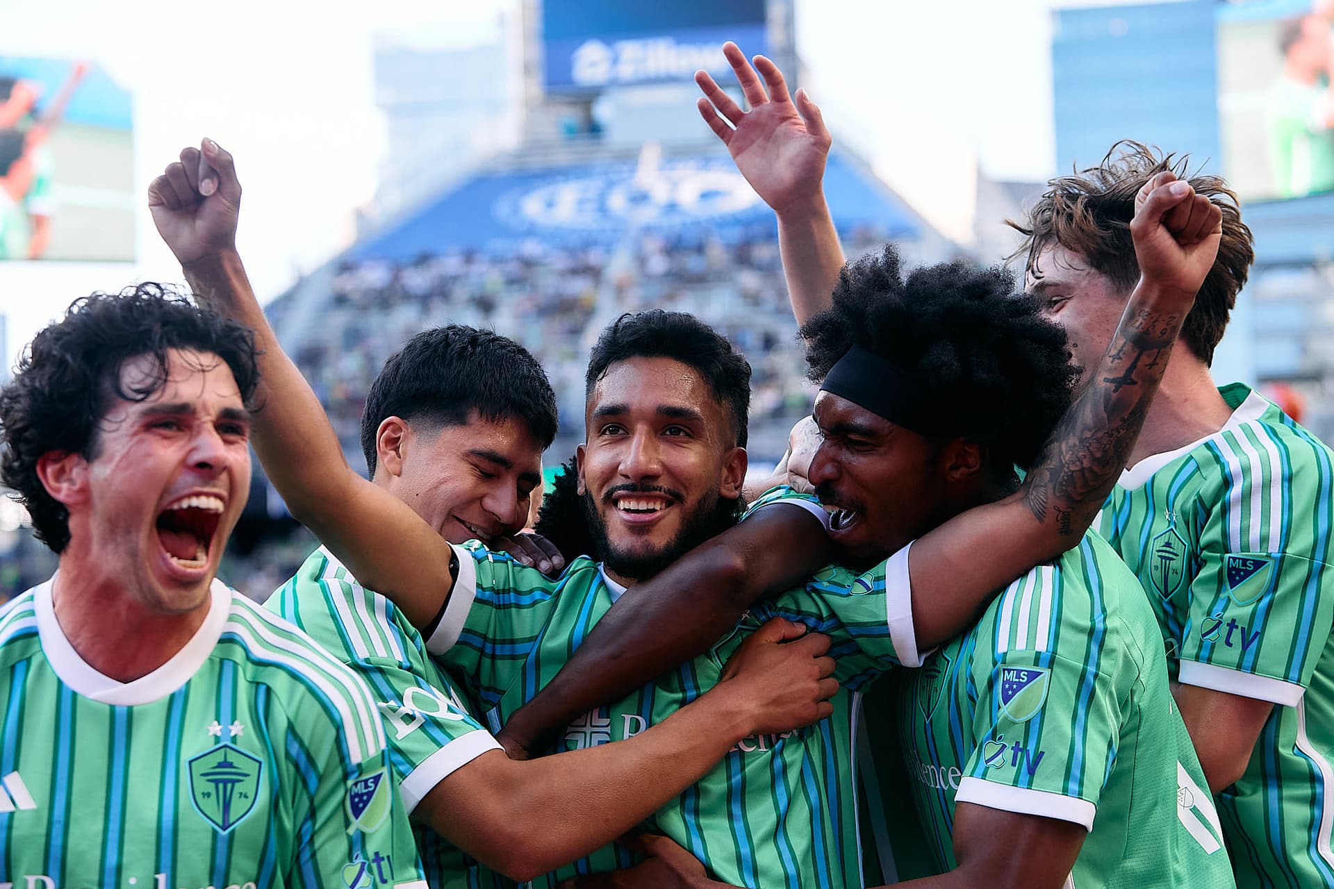 Seattle Sounders FC players celebrating a goal scored by Jesús Ferreira (9) during a Major League Soccer match. The team is gathered around Ferreira, who is in the center of the celebration, with arms raised in triumph. The vibrant green field and the stands filled with cheering fans create an electrifying atmosphere, capturing the excitement of the moment.